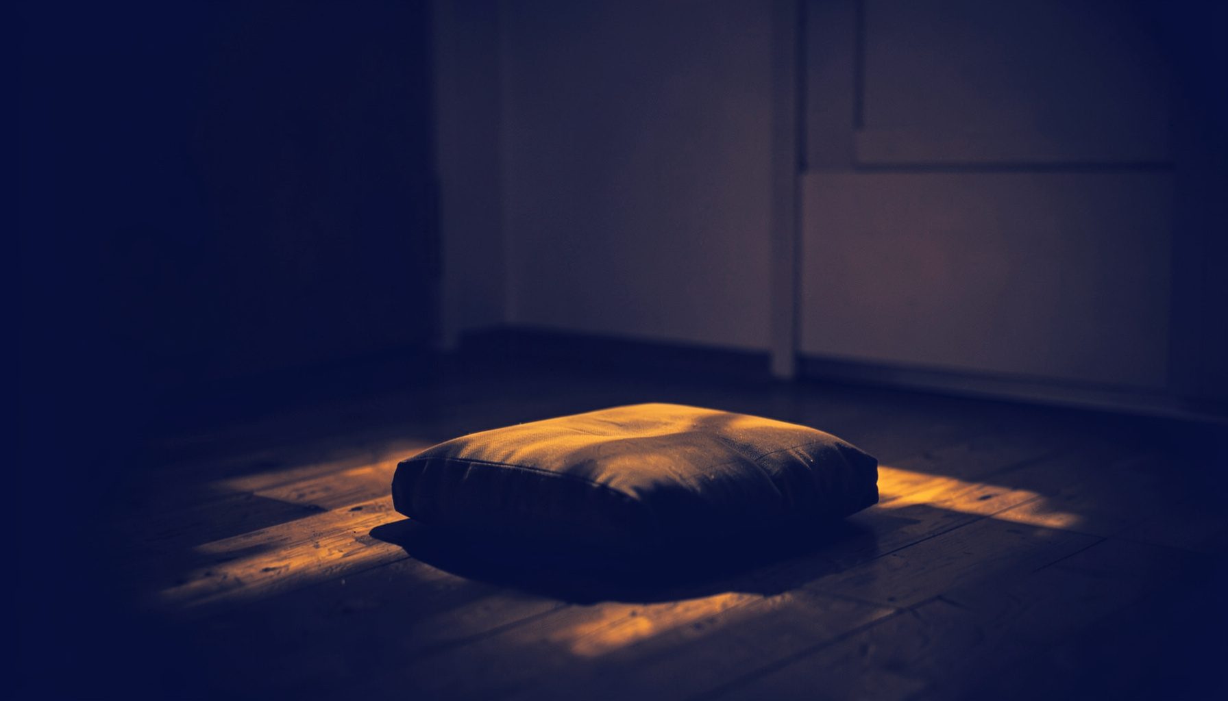 Soft duotone photograph in deep indigo and warm amber tones, showing a dimly lit meditation space with a simple cushion on a wooden floor, gentle candlelight casting long shadows across the room, evoking calm introspection.