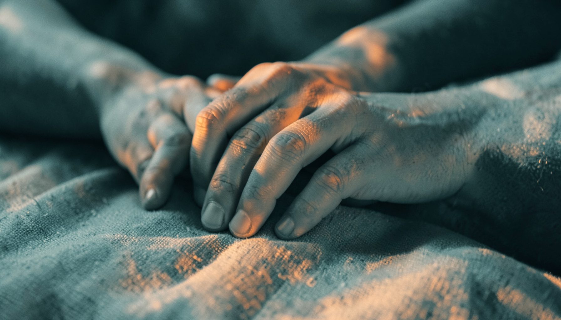 Duotone photograph in muted teal and warm copper, showing a close-up of hands resting gently on a textured fabric, soft natural light falling across the scene, conveying stillness and mindful presence.